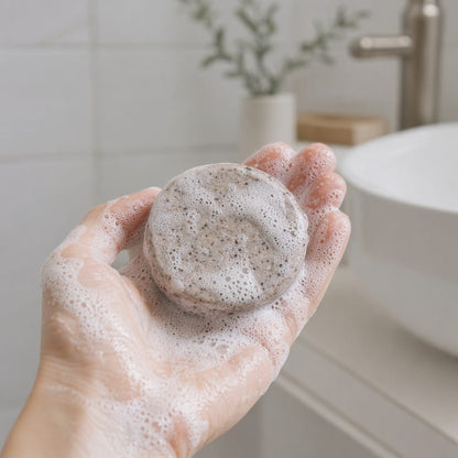 Hand holding a lathered scalp balancing shampoo bar with rich white foam — herbal shampoo for hair loss concerns, shown in a modern Scandinavian bathroom with sink visible in background